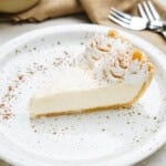 A slice of cream pie on a speckled plate, garnished with whipped cream and cocoa powder. Forks and a bowl are visible in the background.