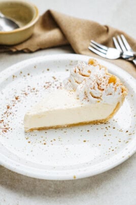 A slice of cream pie on a speckled plate, garnished with whipped cream and cocoa powder. Forks and a bowl are visible in the background.