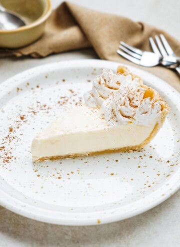 A slice of cream pie on a speckled plate, garnished with whipped cream and cocoa powder. Forks and a bowl are visible in the background.