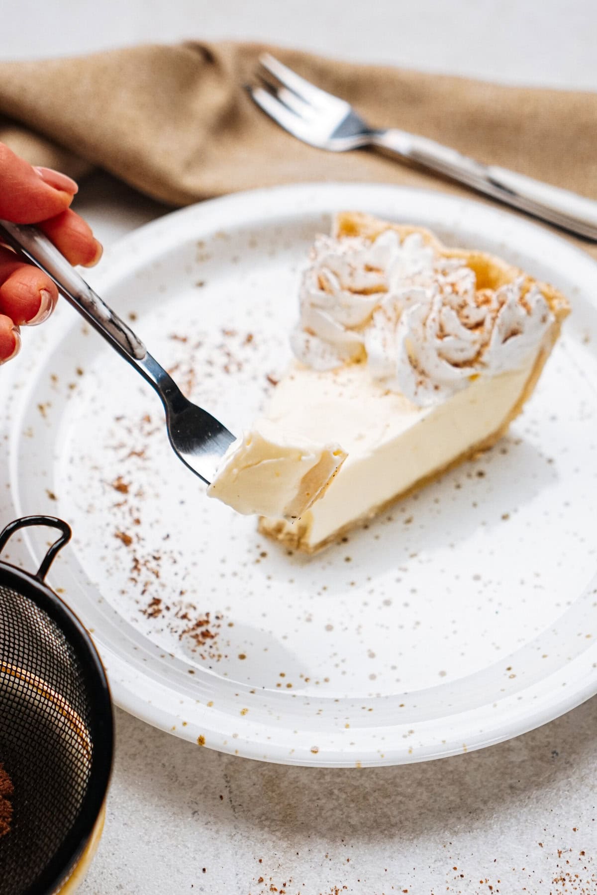 A hand holding a fork with a bite of cream pie on a white plate, garnished with whipped cream and chocolate shavings. A napkin and fork are in the background.