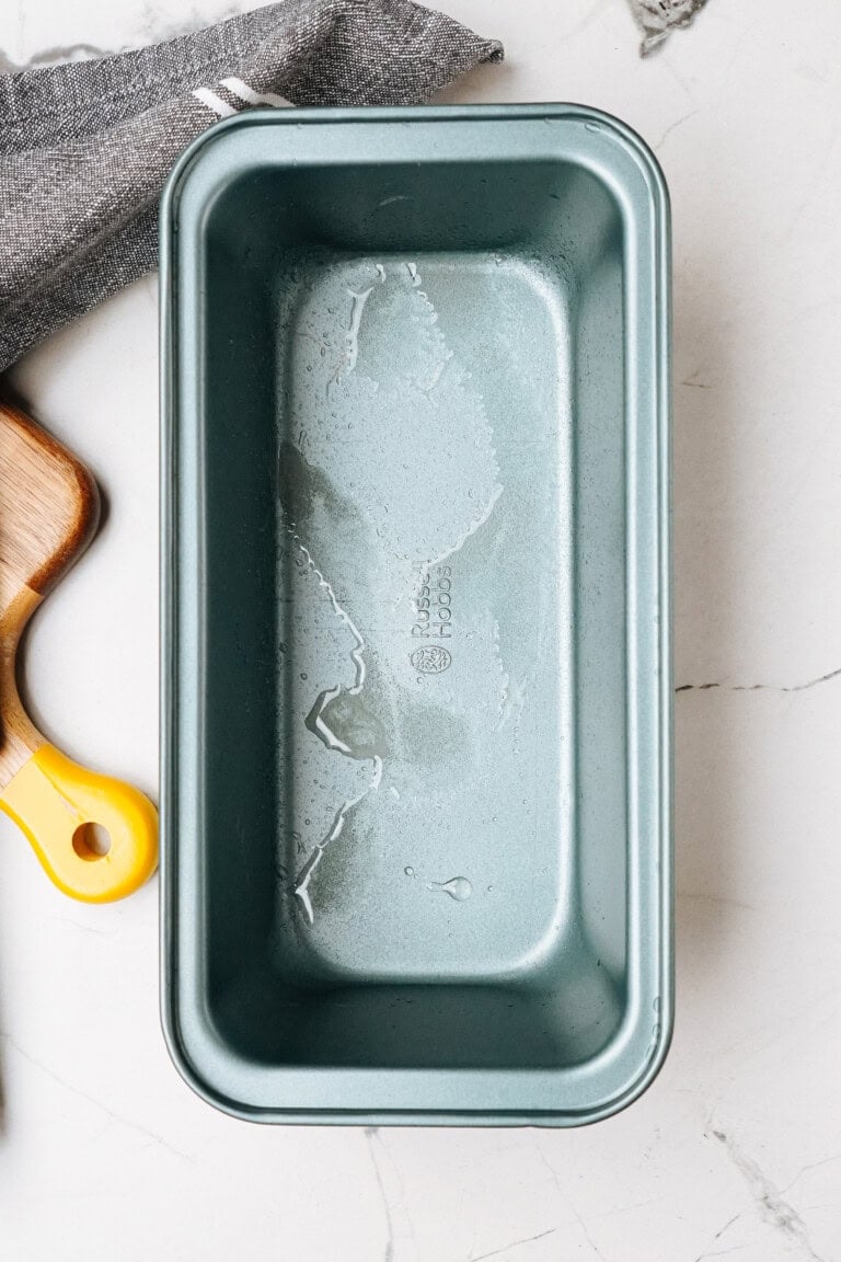 Empty metal loaf pan on a marble surface with a wooden kitchen tool nearby.