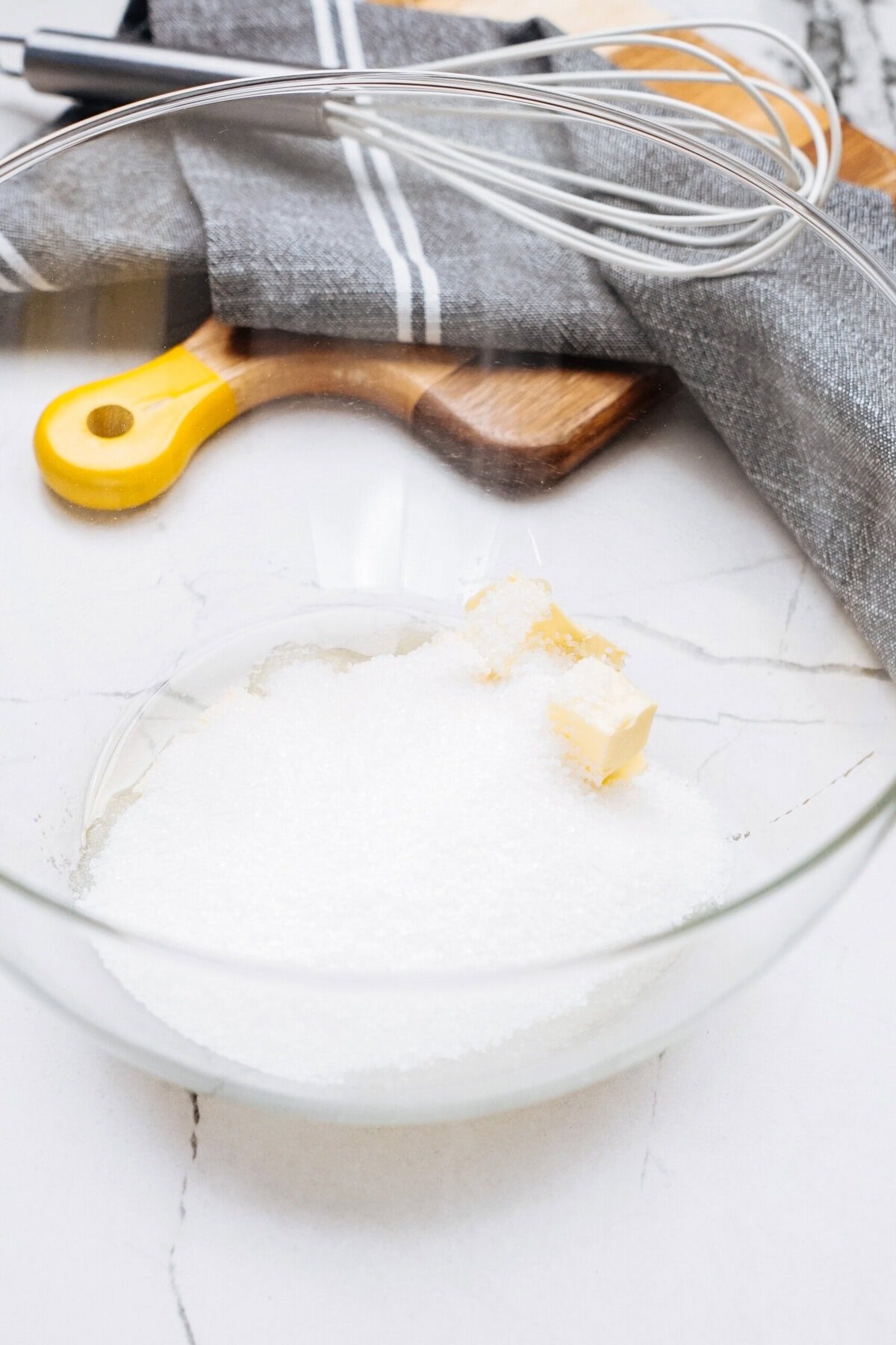 A glass bowl with sugar and butter cubes on a marble countertop, next to a whisk, a wooden cutting board, and a gray cloth.