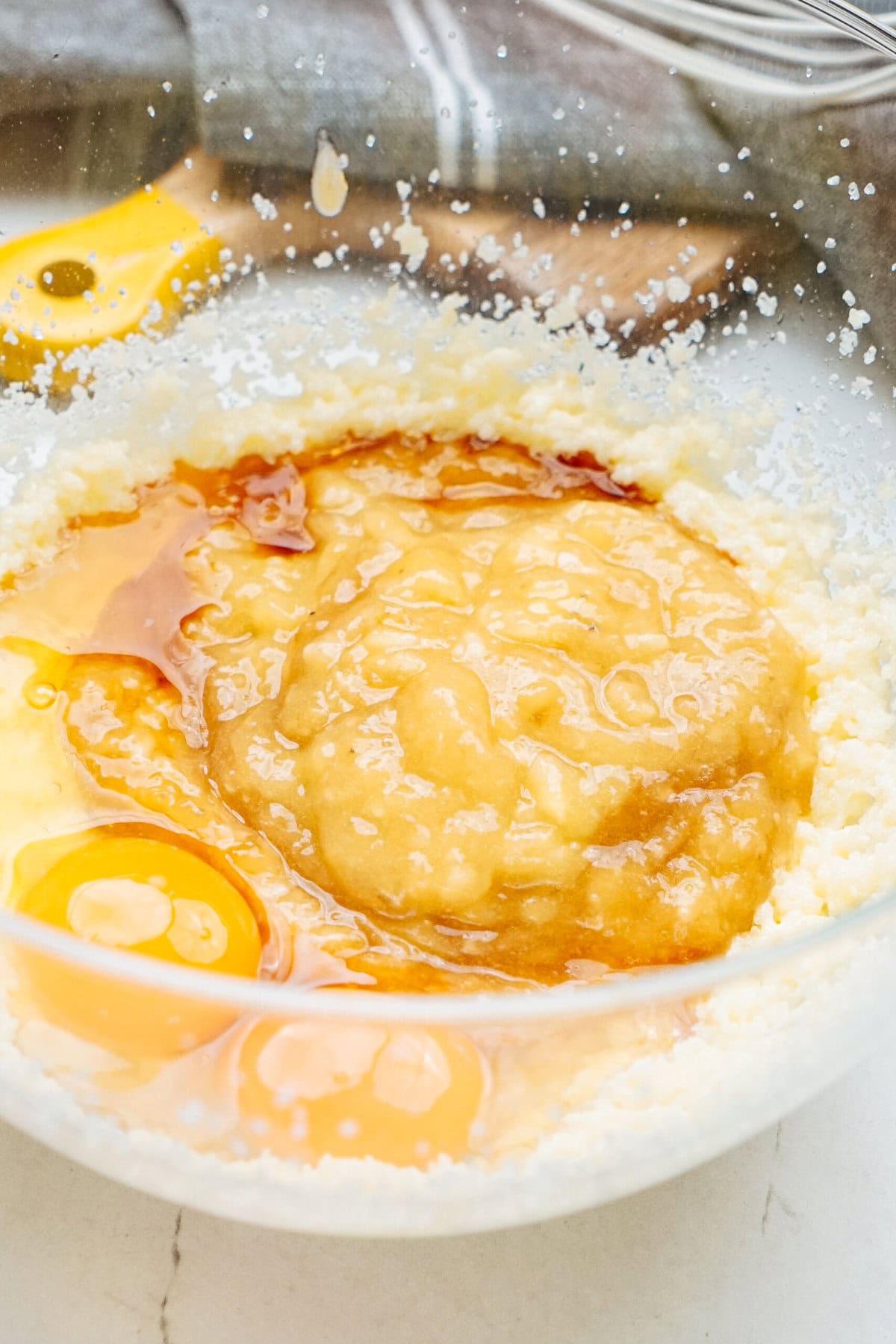 A glass bowl containing a mixture of sugar, butter, banana, and raw eggs on a kitchen counter.