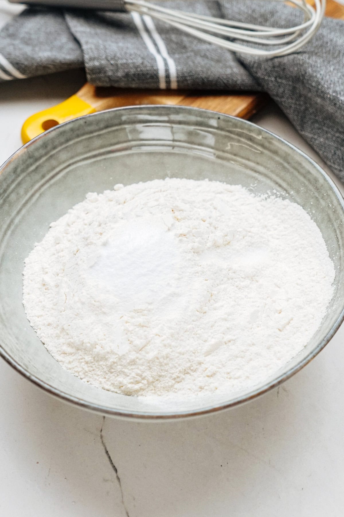 A bowl of white flour with a whisk and cloth on a wooden board in the background.