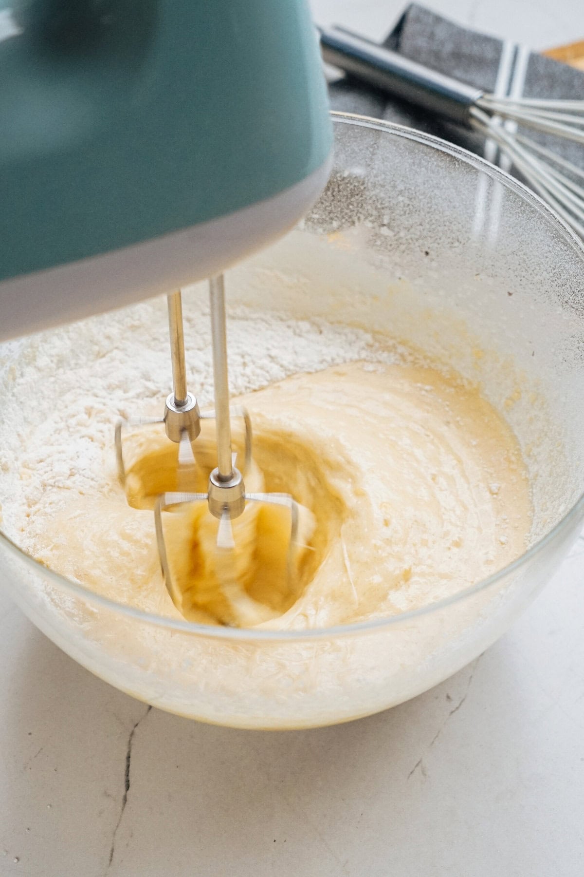 A hand mixer blending a creamy batter in a glass bowl, surrounded by baking utensils on a light surface.