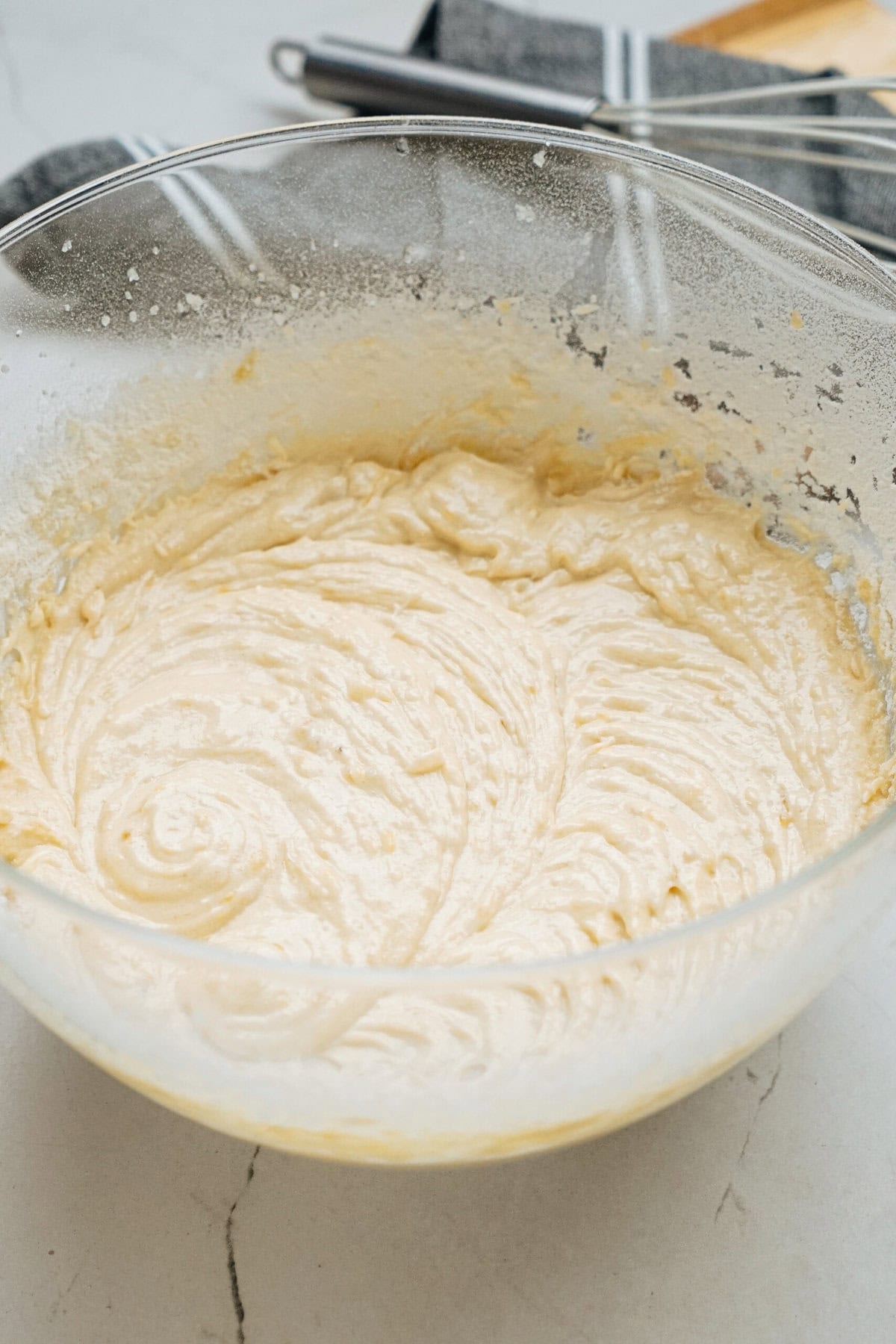 Batter in a clear glass bowl with a whisk and striped cloth in the background on a kitchen countertop.