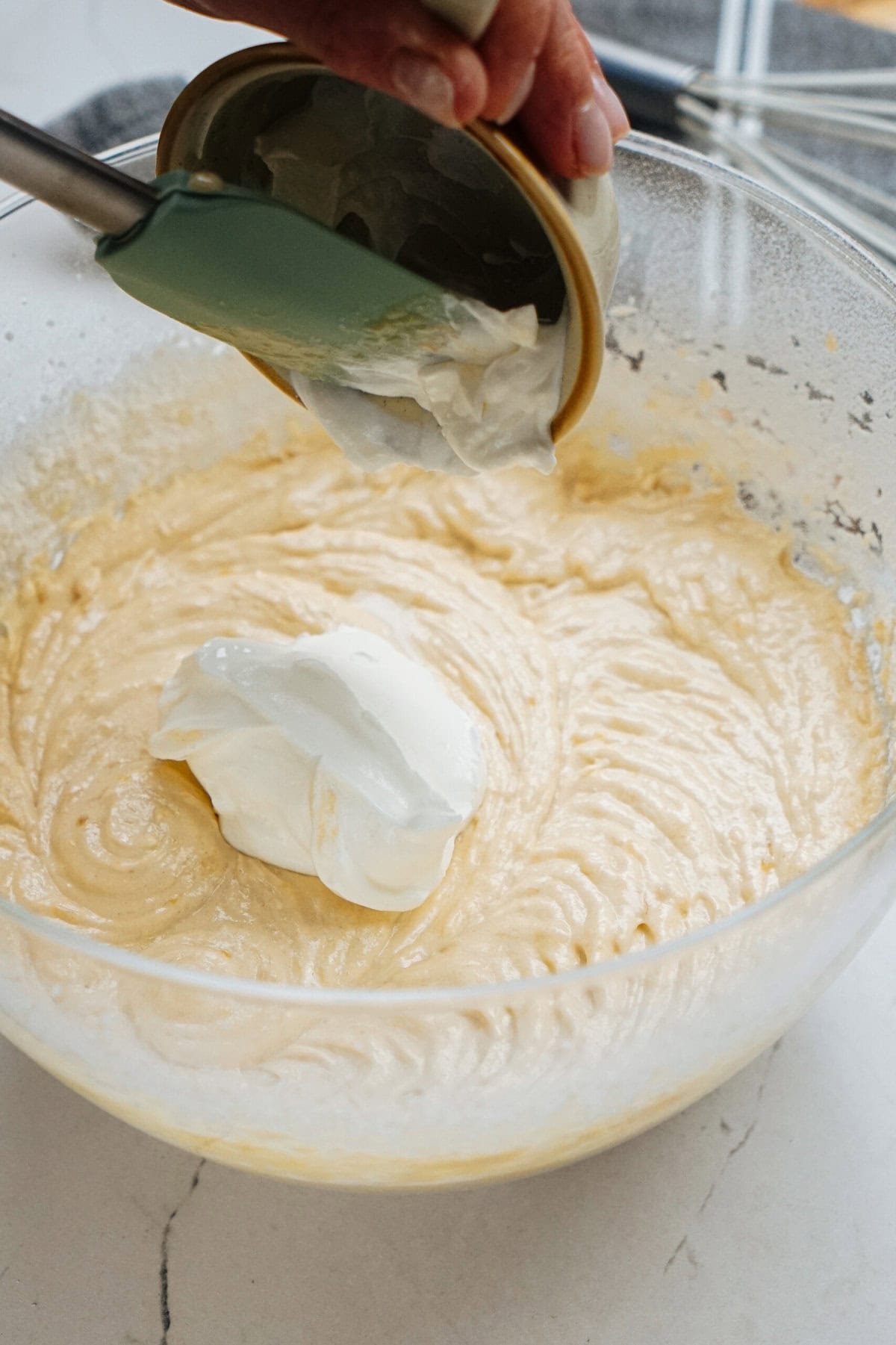 A spatula is used to add sour cream into a bowl of cake batter being mixed.