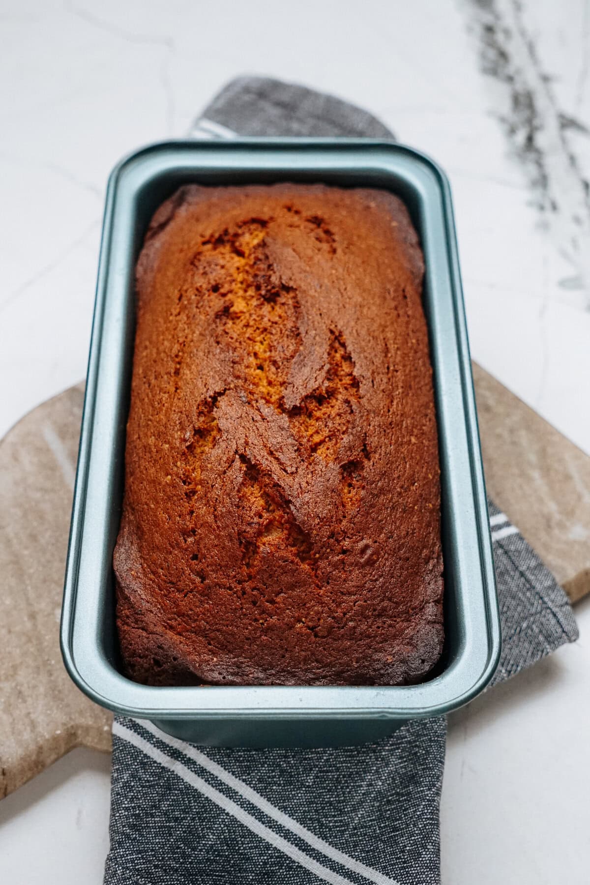 A loaf of banana bread in a metal pan on a marble surface, with a striped cloth underneath.