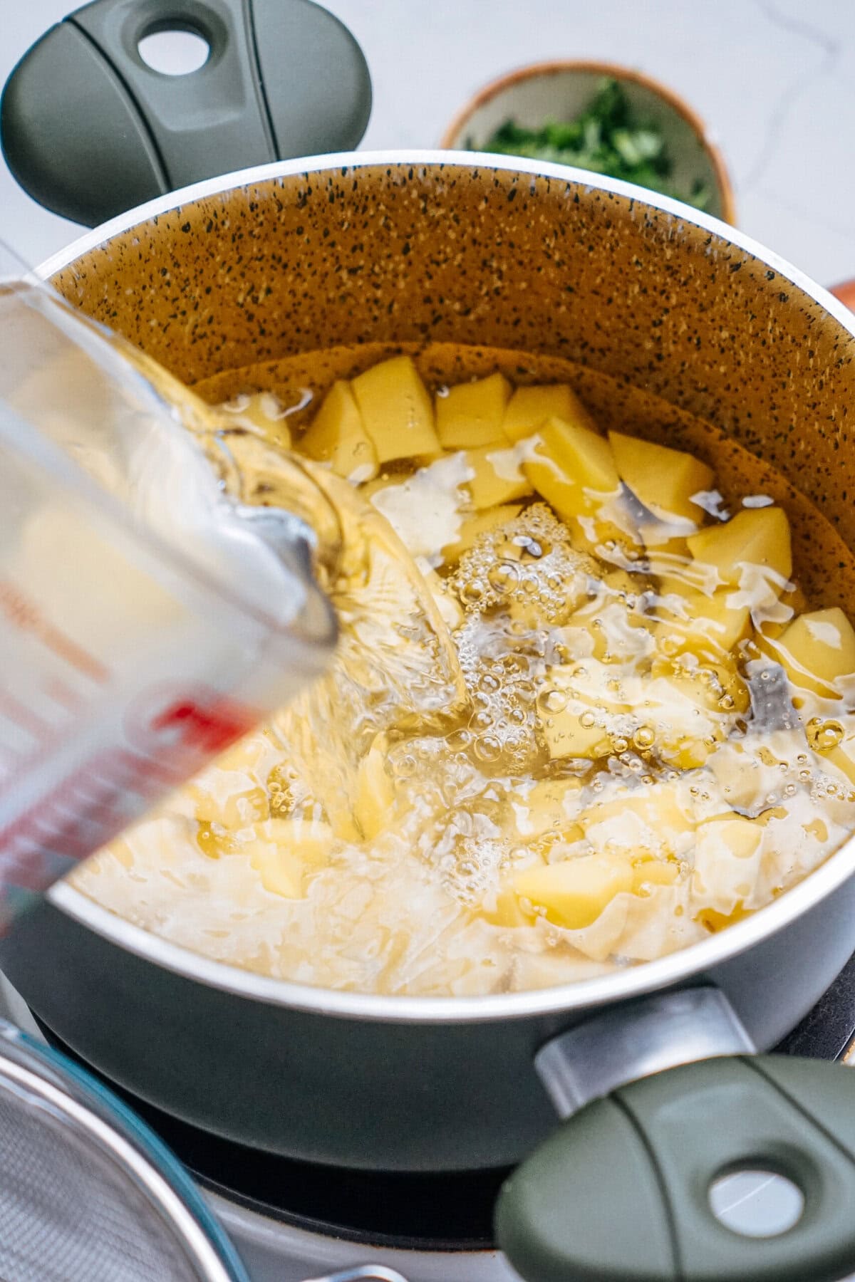 A saucepan with chopped potatoes being boiled. Liquid is being poured from a measuring cup into the pot.