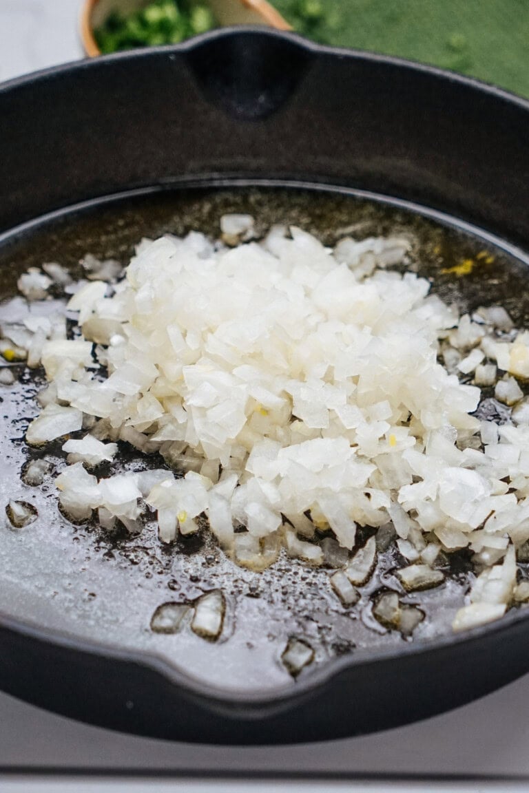 Chopped onions in a cast iron skillet, with a green vegetable blurred in the background.