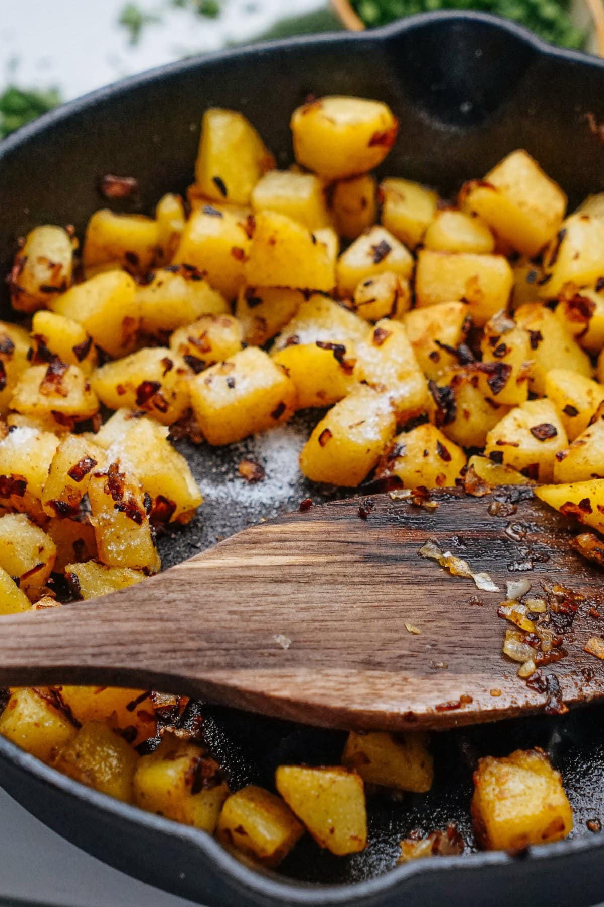 Diced potatoes and onions are being sautéed in a skillet with a wooden spatula.