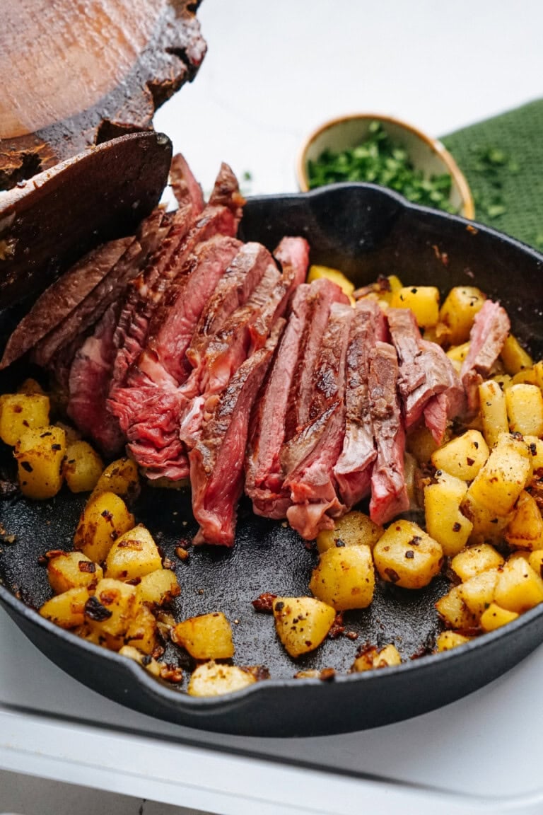 Sliced steak and seasoned potatoes in a skillet, with a small bowl of herbs in the background.