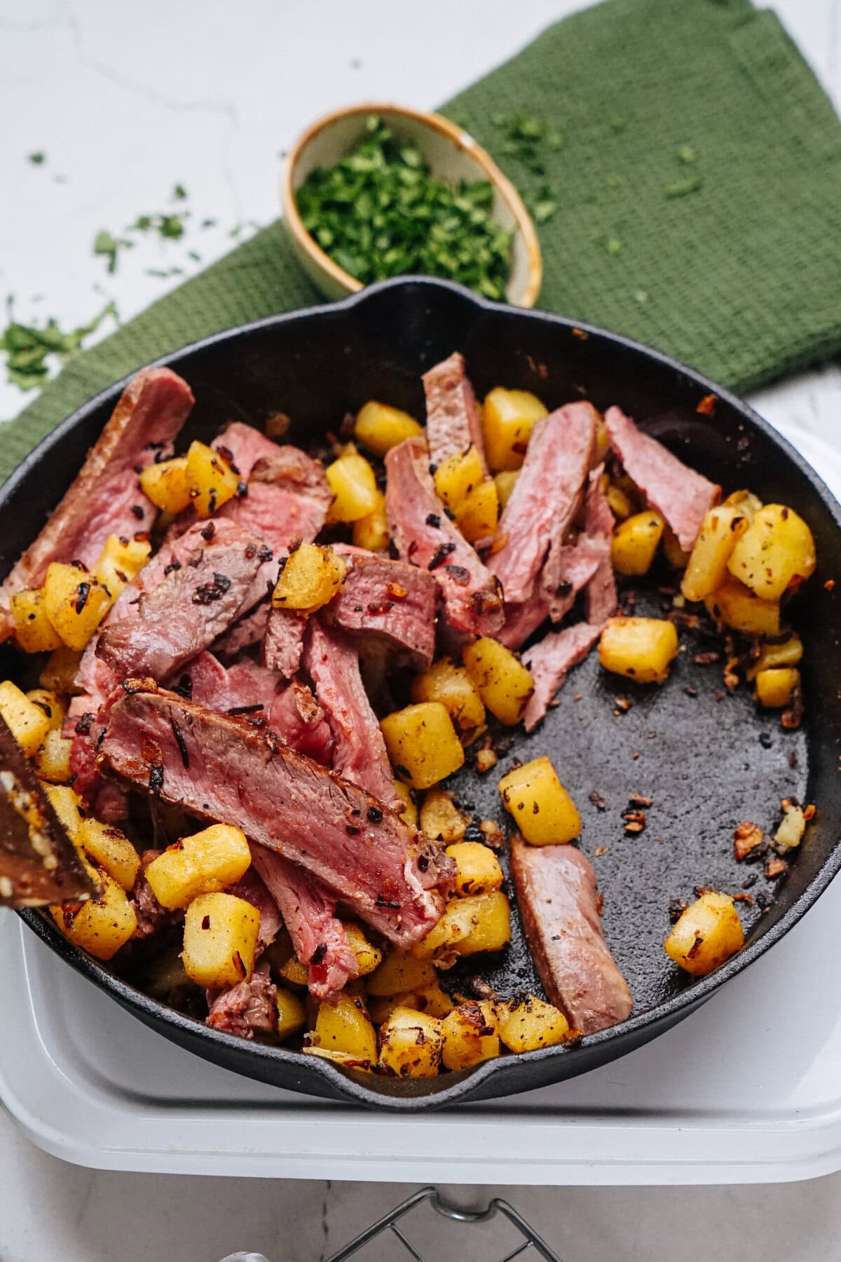 A skillet with sliced steak and cubed potatoes on a white surface, garnished with chopped herbs.