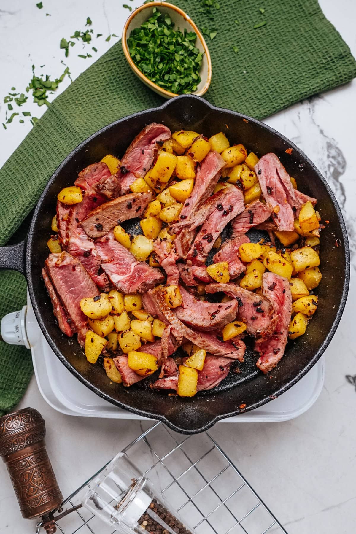 A skillet with sliced corned beef and cubed potatoes on a table, garnished with chopped herbs.