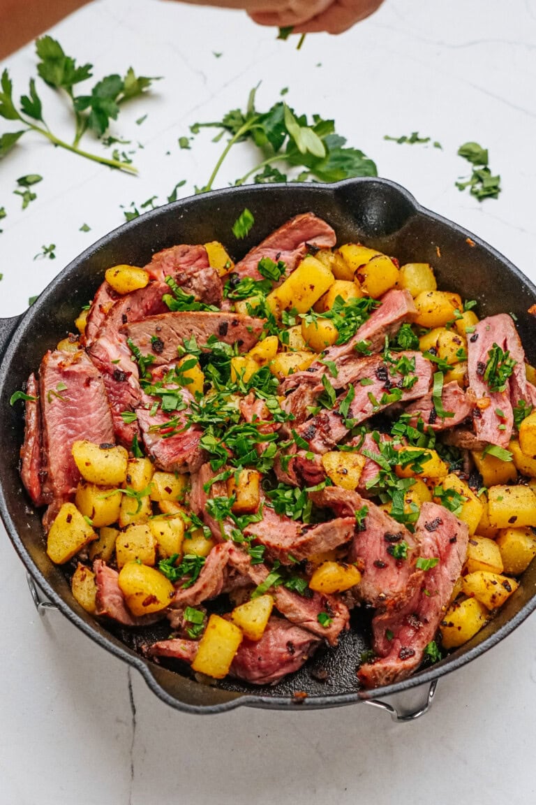 A skillet filled with sliced cooked meat and roasted potatoes, garnished with chopped fresh parsley on a white surface.