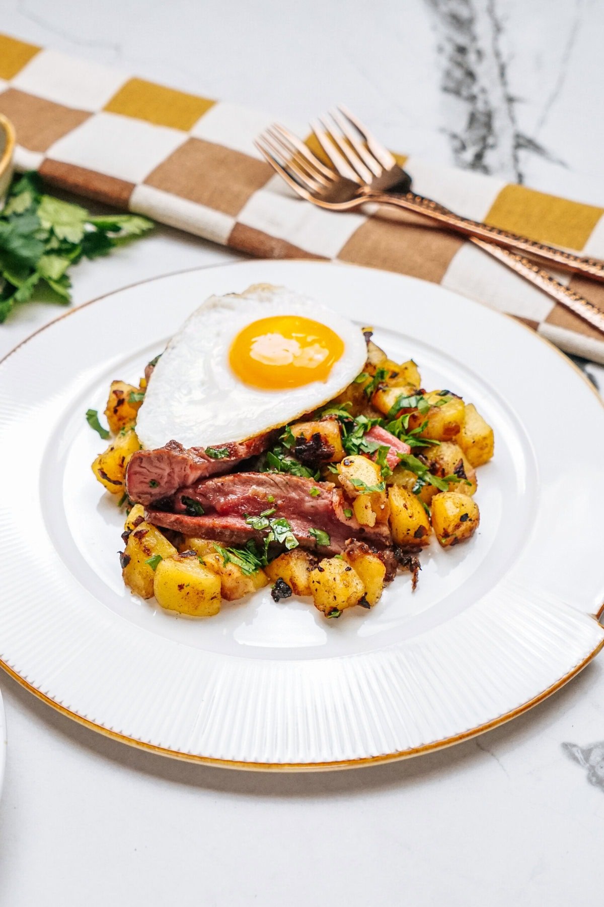 A plated dish with sliced steak, roasted potatoes, and a sunny-side-up egg on top, garnished with green herbs.