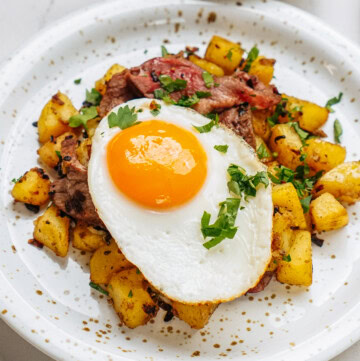 A plate of fried potatoes topped with chopped herbs, a slice of cooked meat, and a sunny-side-up egg. A small bowl sits in the background.