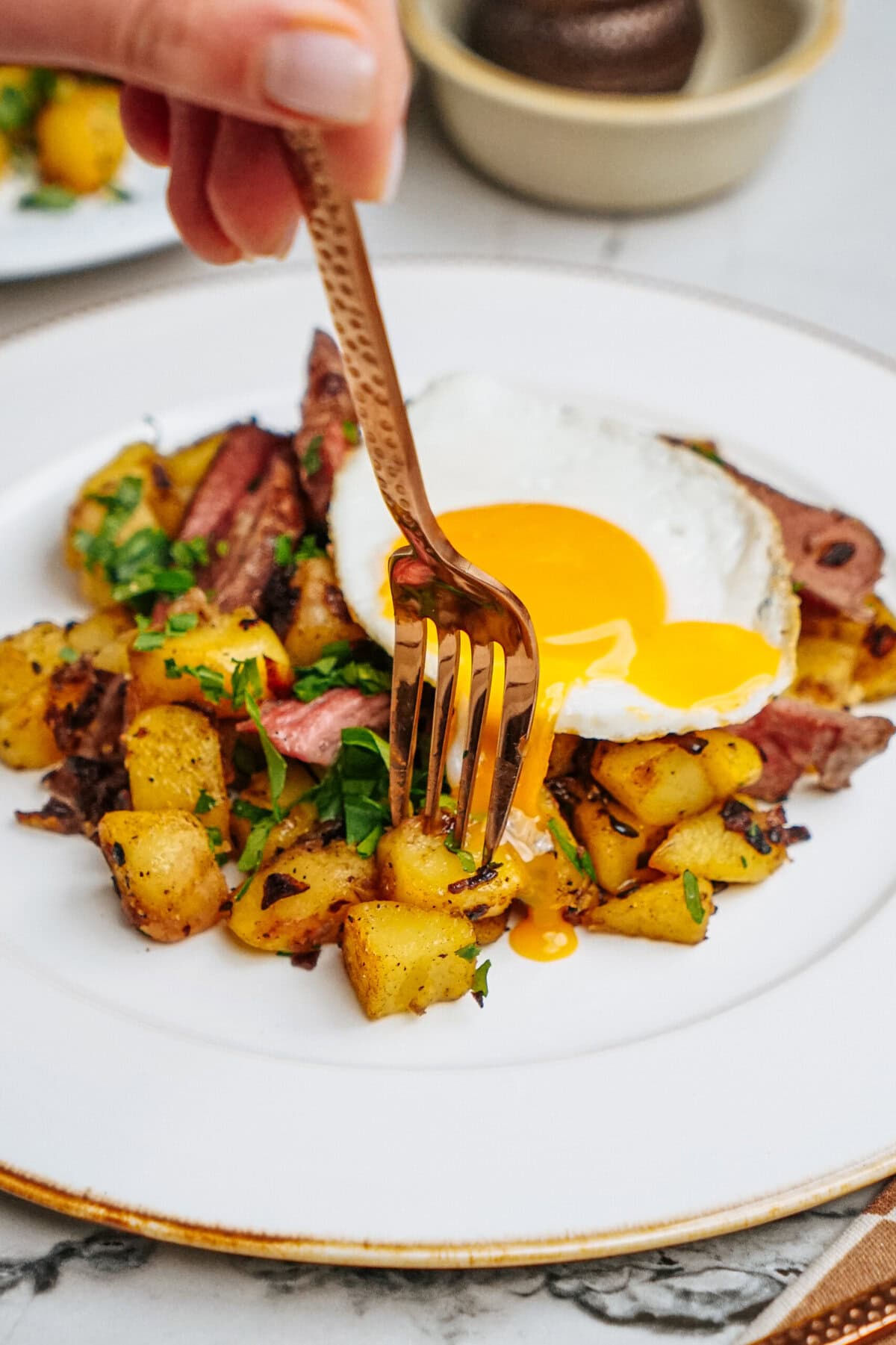 A hand with a fork is about to pierce a fried egg yolk on a plate of diced potatoes topped with herbs.