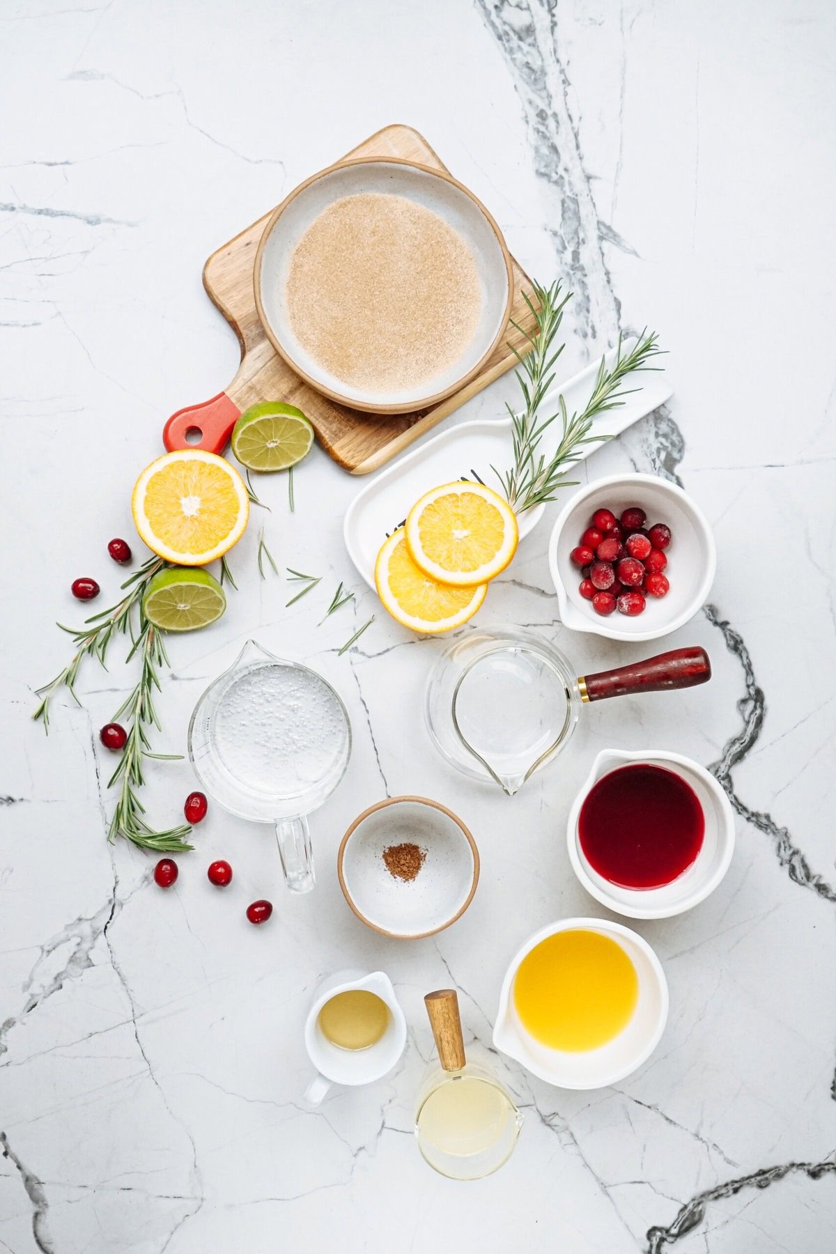 Assorted ingredients arranged on a marble surface, including citrus slices, cranberries, liquid in bowls, and spices, with rosemary as garnish.