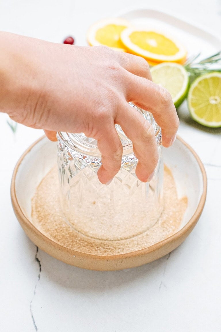 A hand dips a glass rim into a dish of sugar. Slices of orange, lime, and a sprig of rosemary are on a plate in the background.