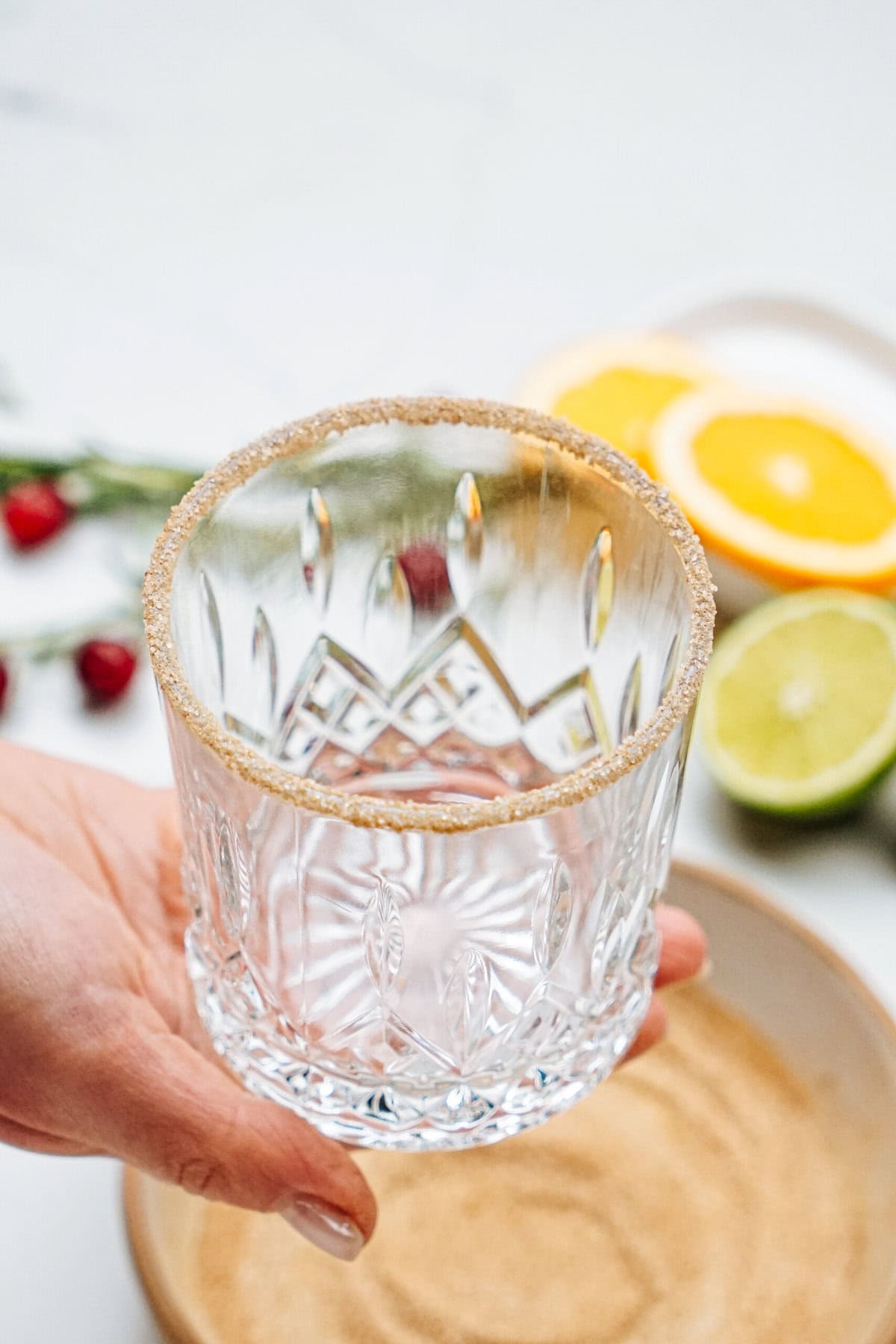 A hand holding a crystal glass with a sugar rim, surrounded by sliced citrus fruits in the background.