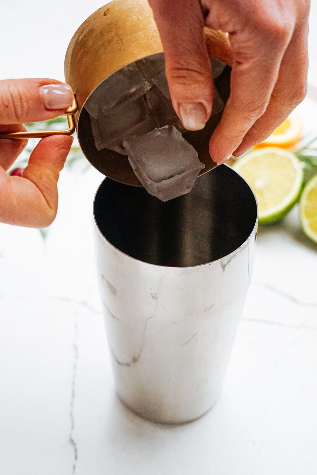 Hands pouring ice cubes from a gold measuring cup into a stainless steel shaker, with sliced limes and an orange in the background on a marble surface.