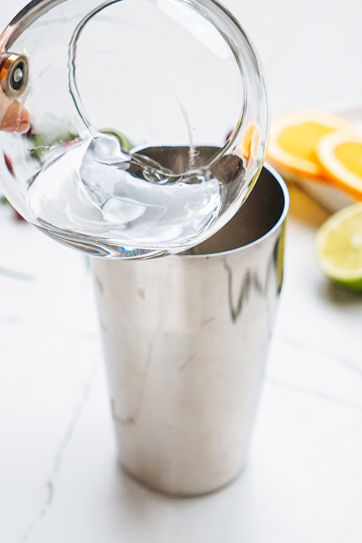 A person pours clear liquid into a stainless steel cocktail shaker. Sliced citrus fruits are visible in the background.