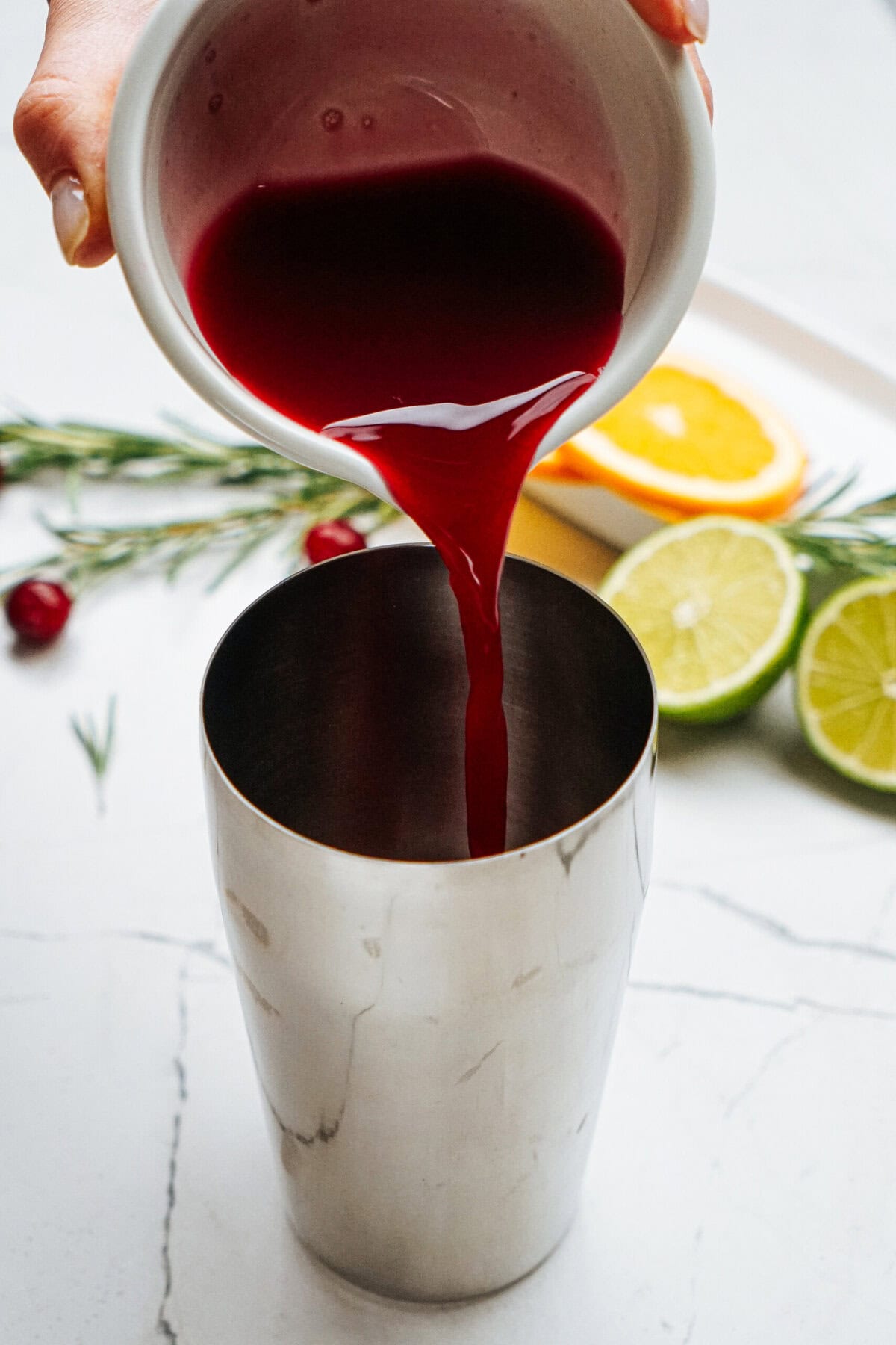 Red liquid being poured into a stainless steel cocktail shaker, with slices of lime and orange, rosemary, and cherries in the background on a marble surface.