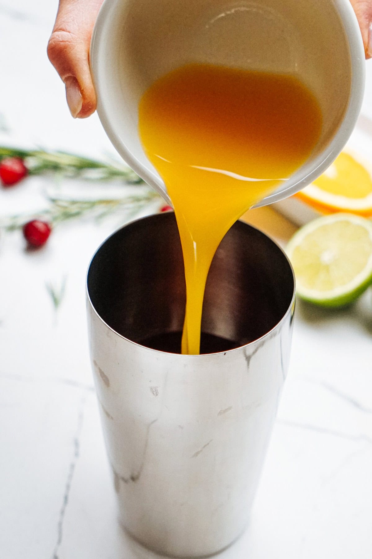 A hand pours orange juice from a small white bowl into a metal shaker on a marble countertop with sprigs of rosemary, cranberries, and sliced citrus in the background.