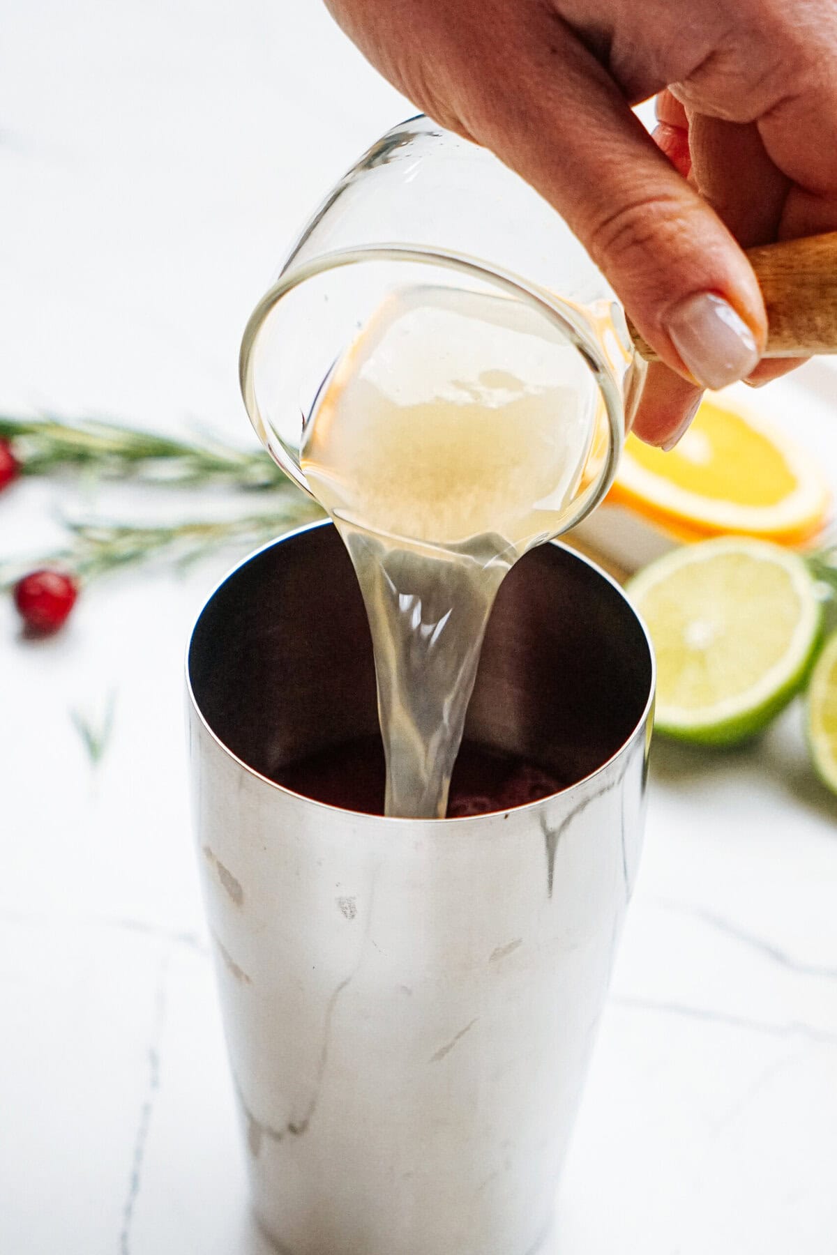 A hand pours liquid from a clear cup into a stainless steel shaker. In the background, there are sliced citrus fruits and a sprig of rosemary on a white surface.