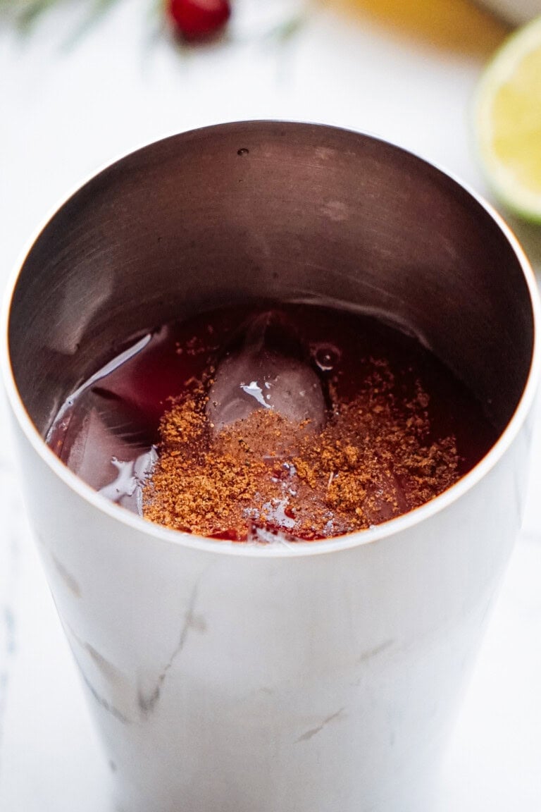 A stainless steel cup filled with a dark red liquid and ice, topped with a sprinkle of brown spice. A lime wedge is visible in the background.