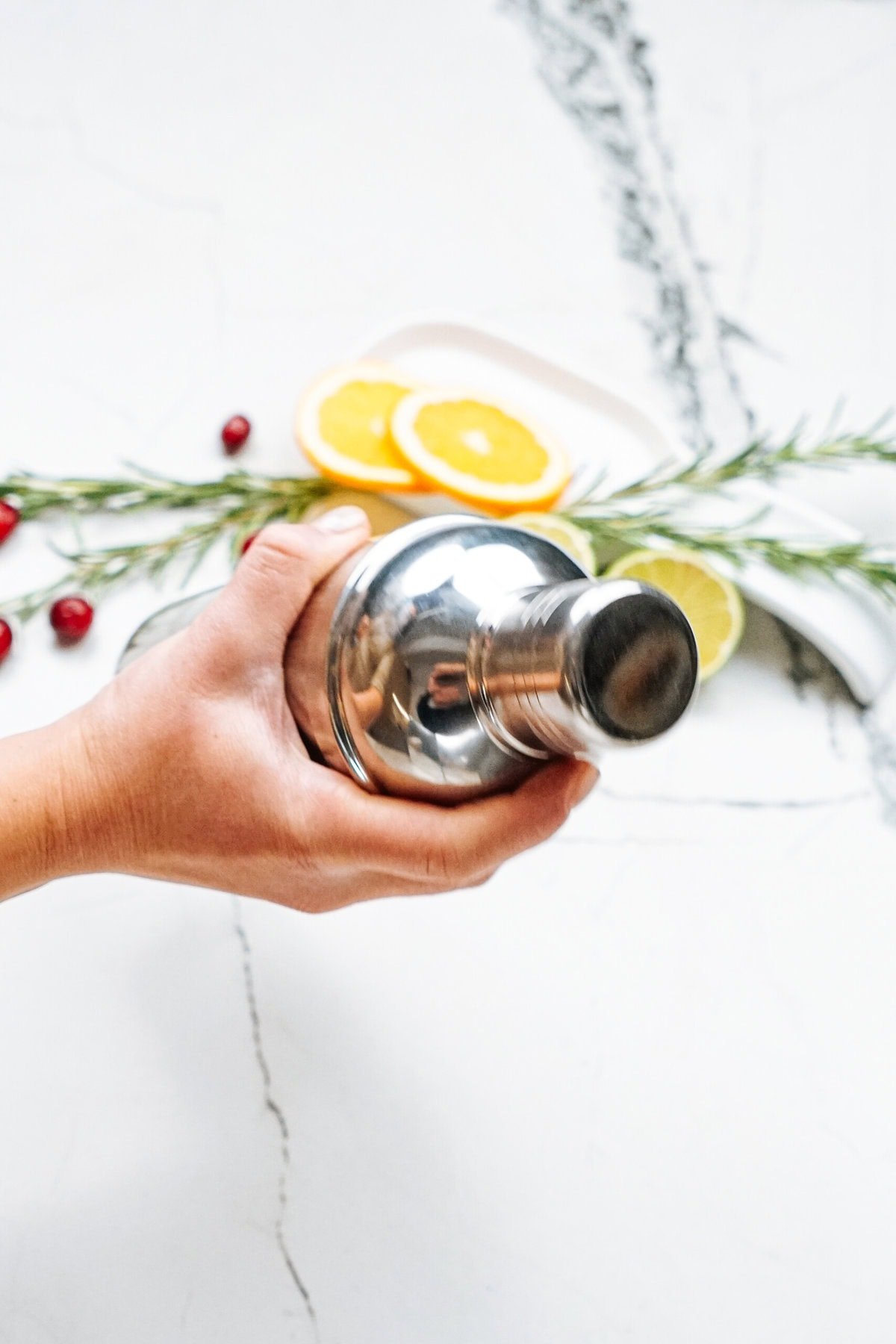 A hand holding a stainless steel cocktail shaker over a marble surface, with sliced lemons and oranges, cranberries, and rosemary in the background.