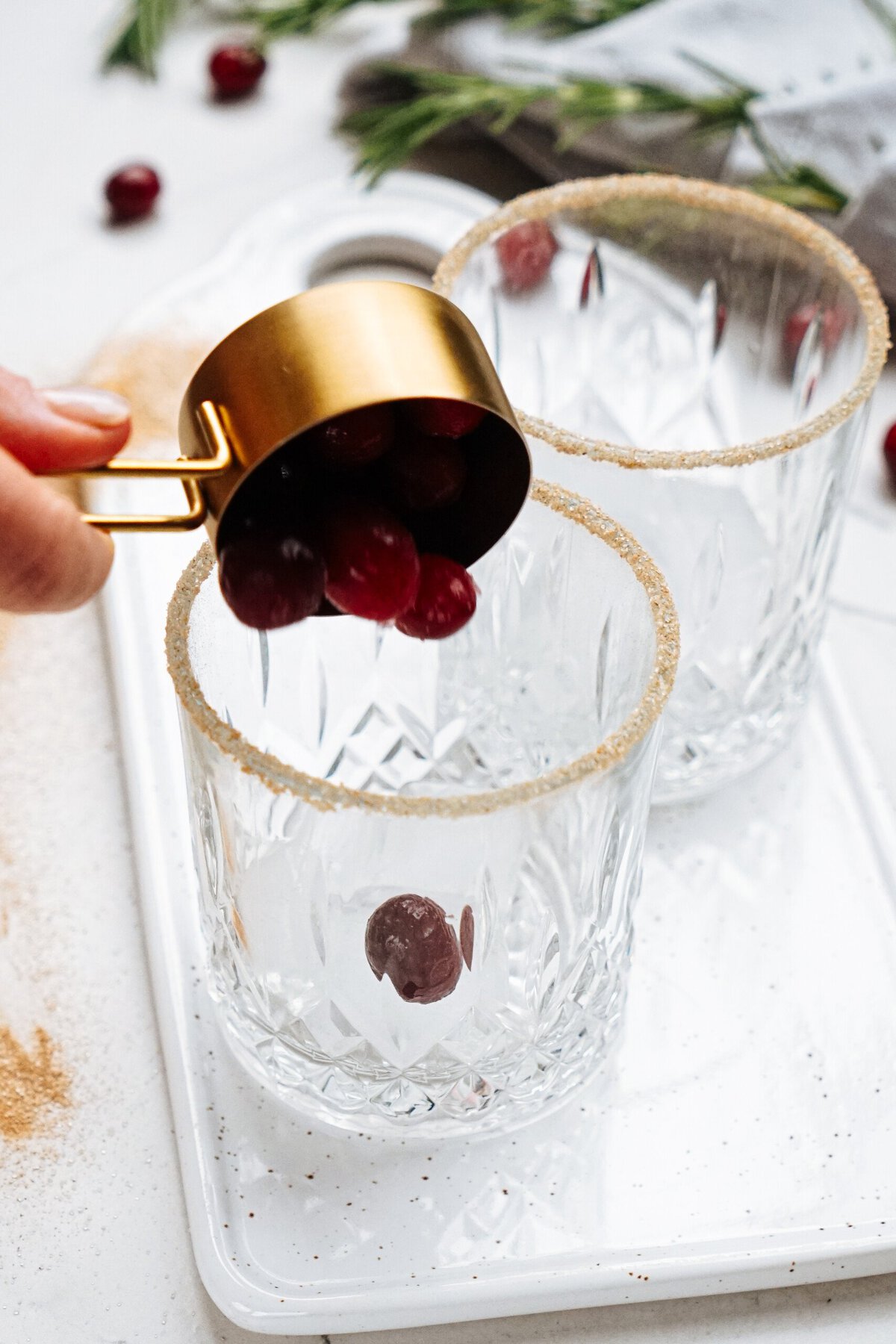 Hand dropping cranberries into crystal glasses with sugared rims on a white tray.