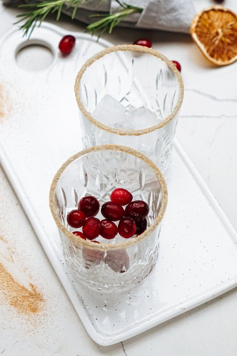 Two empty glasses with sugar rims sit on a tray, one containing ice and cranberries, surrounded by scattered cranberries and rosemary.