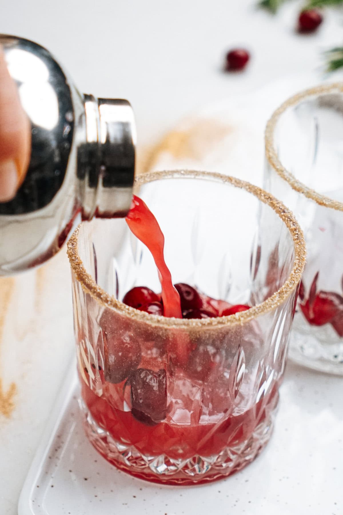 Pouring red cocktail from shaker into a glass with ice and cranberries, rimmed with sugar.