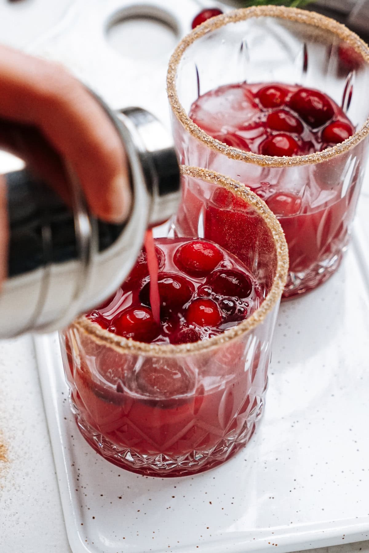 Close-up of two glasses filled with cranberry cocktails, each garnished with whole cranberries and sugar-rimmed edges. A hand pours liquid from a shaker into one of the glasses.