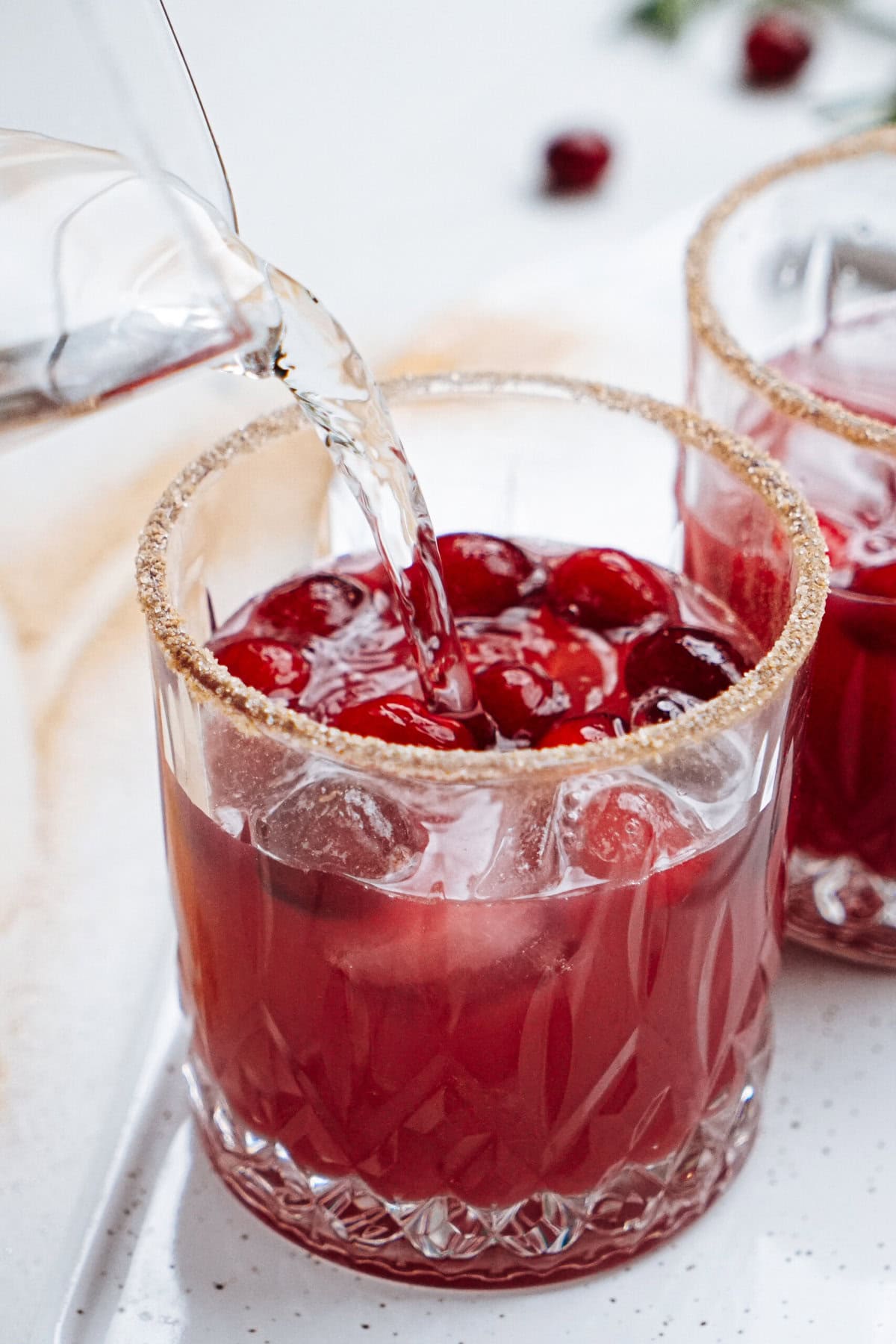 A glass of red beverage with ice and cranberries is being poured, the rim is coated with a sugar mix.