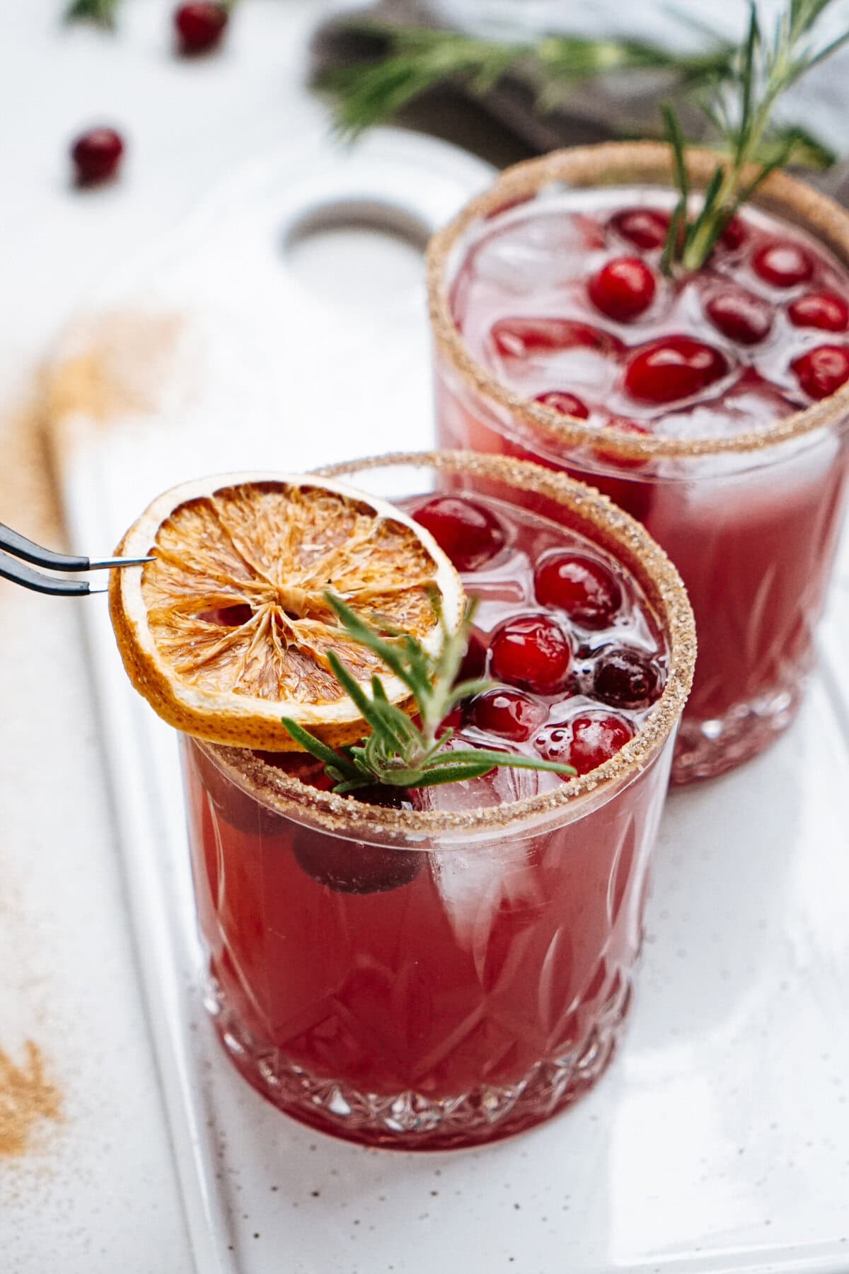 Two glasses of red punch garnished with cranberries, a slice of dried orange, and rosemary, on a white tray.