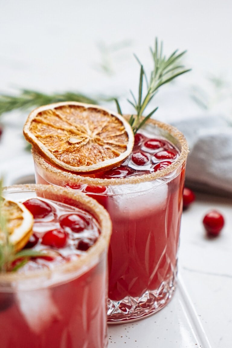 Two glasses of cranberry cocktail, garnished with rosemary and dried orange slices.