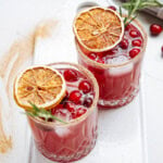 Two glasses of red cocktail garnished with cranberries, dried orange slices, and rosemary, on a white tray.