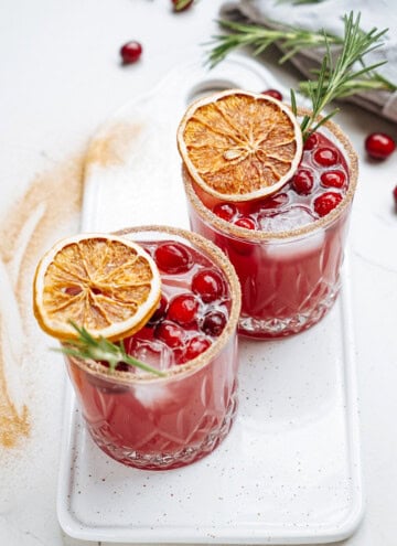 Two glasses of red cocktail garnished with cranberries, dried orange slices, and rosemary, on a white tray.