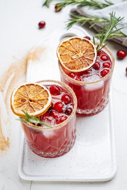 Two glasses of red cocktail garnished with cranberries, dried orange slices, and rosemary, on a white tray.