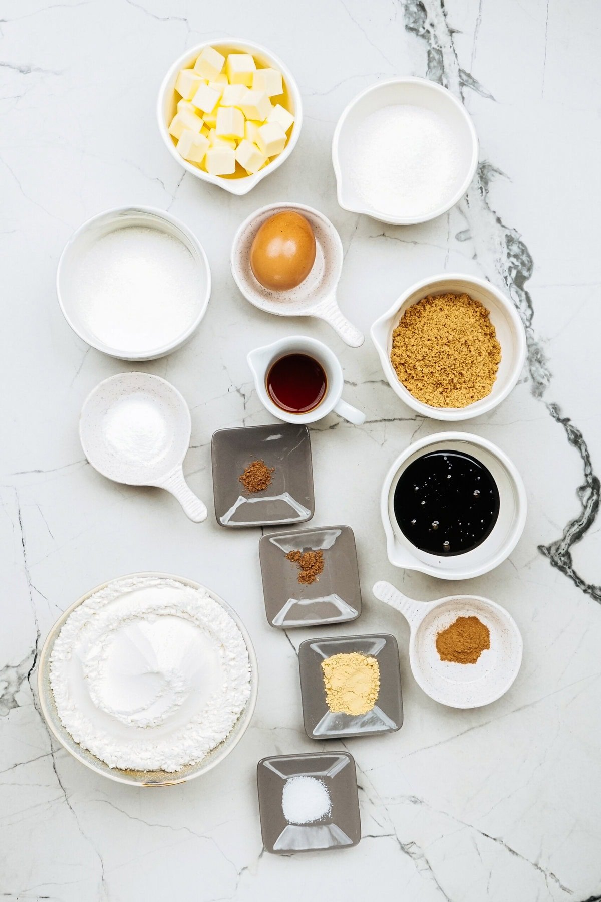 Various baking ingredients for ginger cookies, including butter, sugar, brown sugar, an egg, vanilla extract, molasses, and spices, are arranged on a marble countertop.