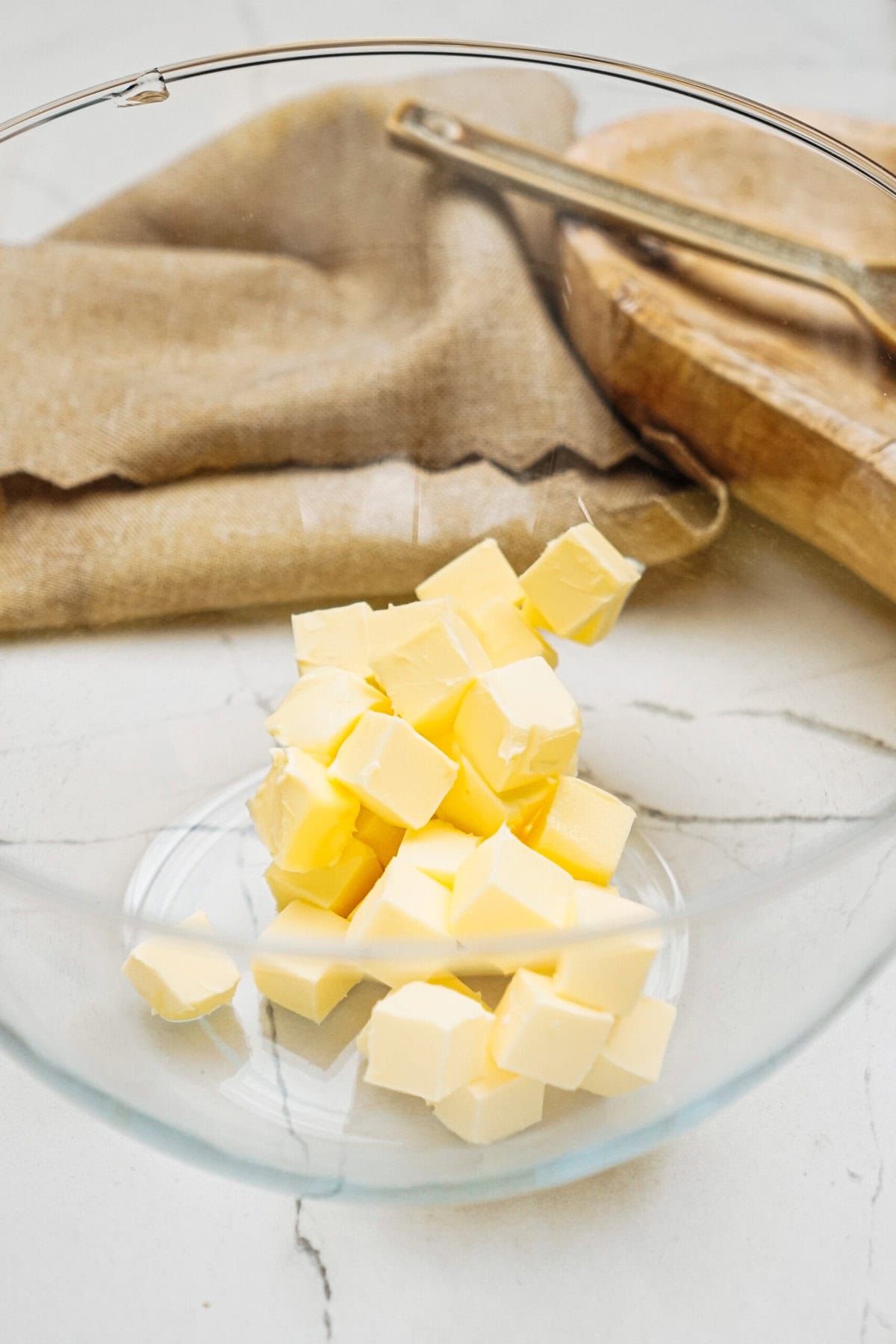 Cubes of butter in a glass bowl on a marble surface, ready to melt into delicious ginger cookies, with a wooden board and beige cloth in the background.