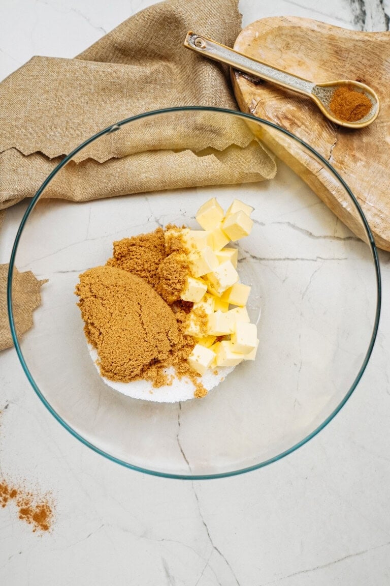 A glass bowl with brown sugar, white sugar, and cubes of butter sits on a marble surface, ready for making ginger cookies. A wooden board holds a spoon with brown powder, and a cloth napkin is nearby.