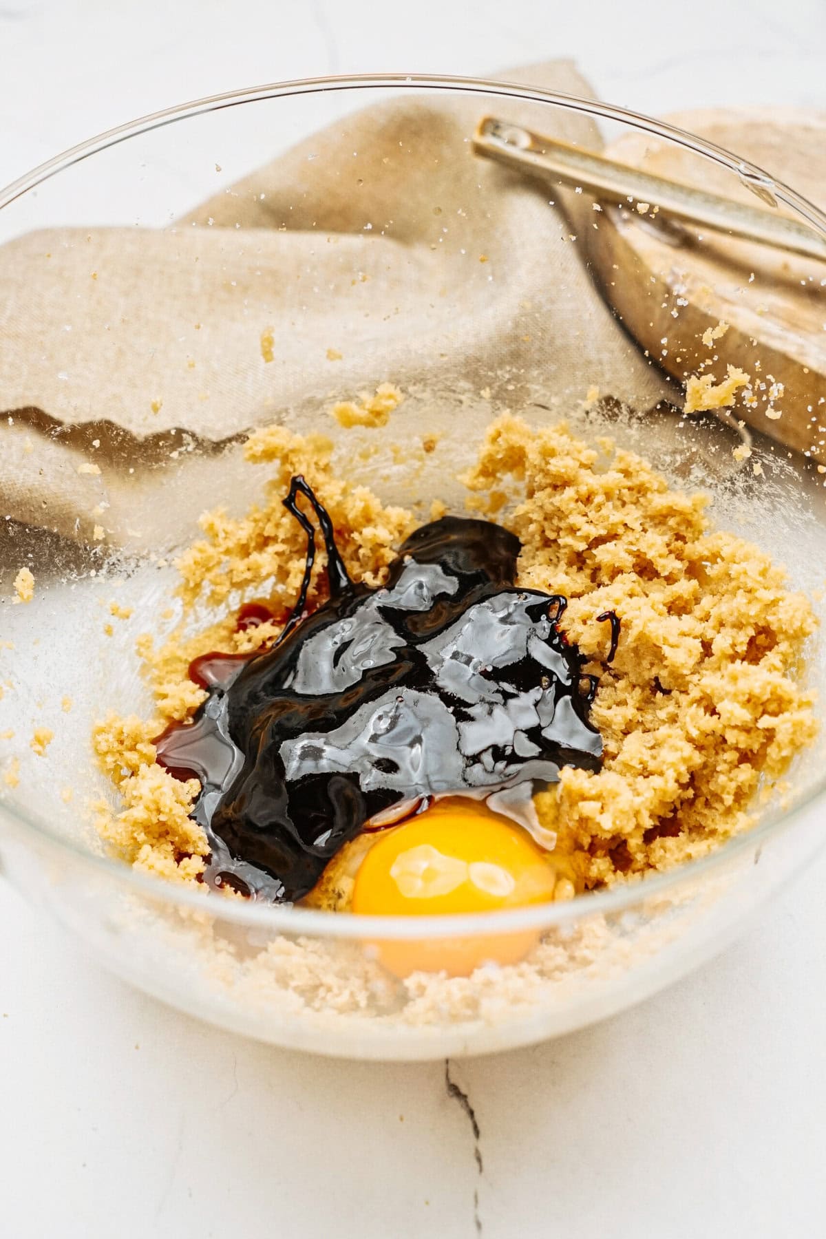 A bowl brimming with cookie dough, an egg, and molasses sits invitingly, ready to transform into delightful ginger cookies. Nearby, a whisk waits patiently on a backdrop of rustic fabric.