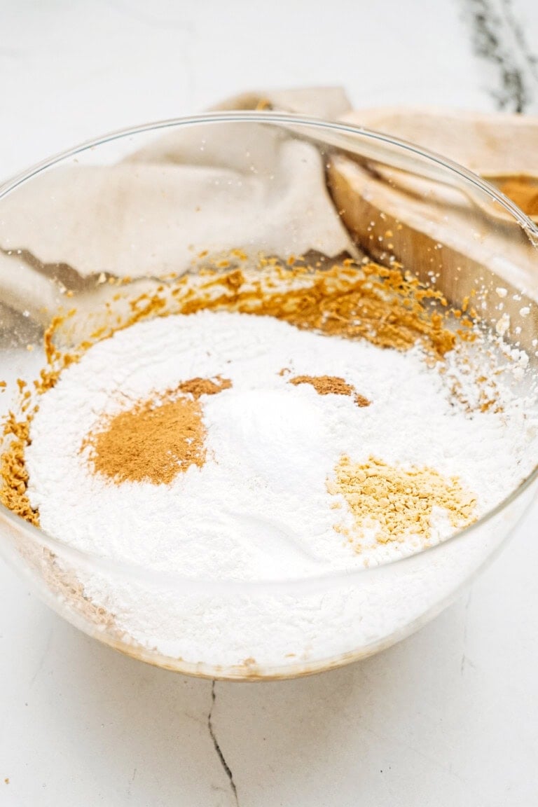 A glass bowl containing flour, baking powder, and spices sits on a white surface, ready to create the perfect ginger cookies.