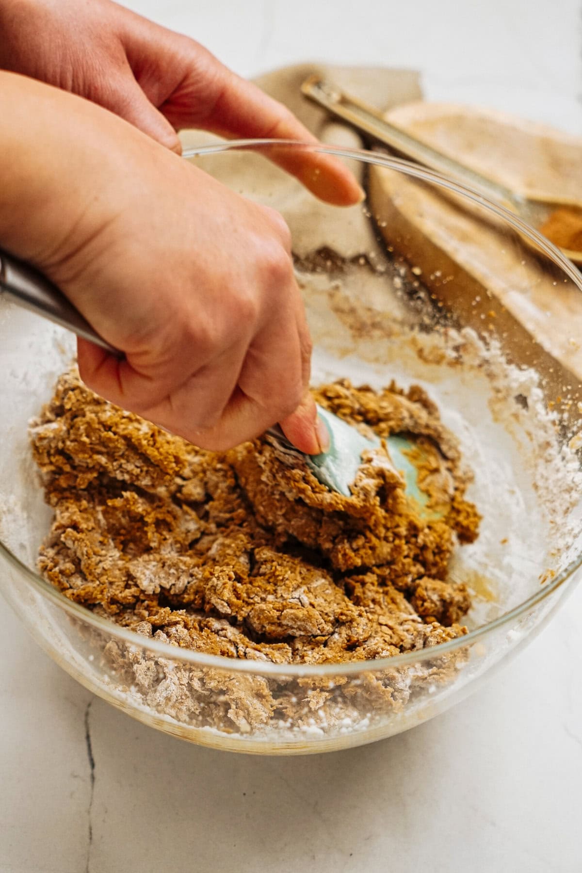 Hands mixing dough for ginger cookies in a glass bowl using a spatula, with a countertop and baking utensils in the background.