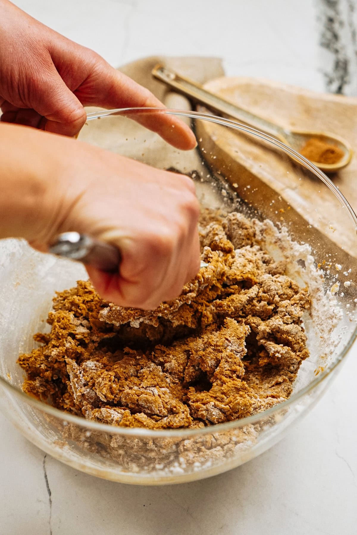 Hands mixing dough in a glass bowl with a metal utensil, crafting the perfect ginger cookies. Nearby are a wooden spoon and a scoop on fabric.