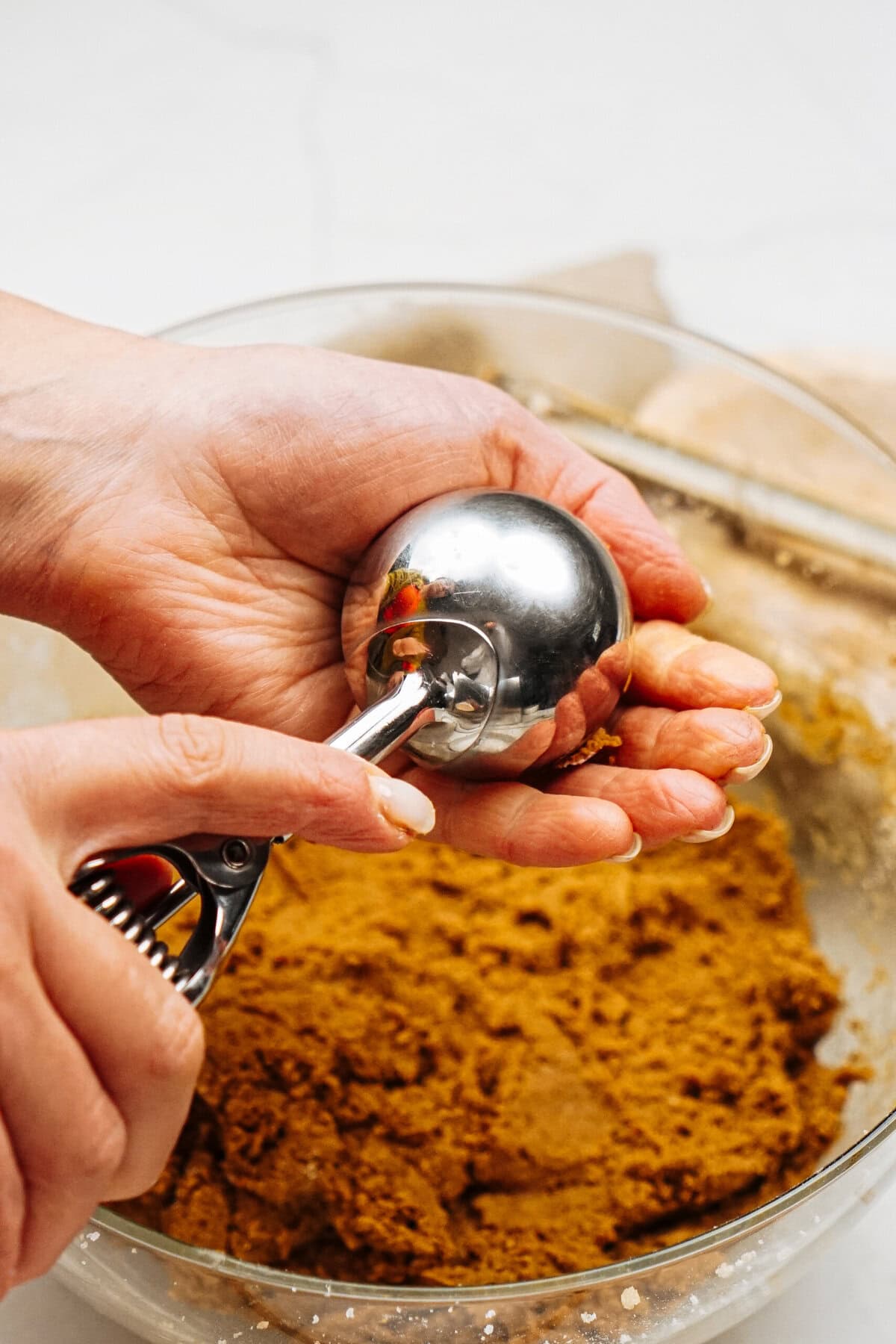 Hands holding a metal cookie scoop over a bowl of brown dough.